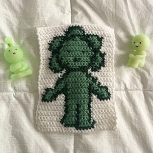 May include: A green crocheted figure of a person with a white background. The figure is standing with its arms outstretched.