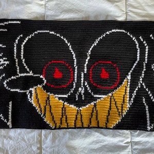 May include: A black and white crocheted pillow with a yellow and red face. The face has two red eyes with a yellow flame inside each eye and a yellow smile.