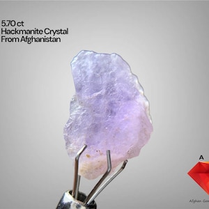 Puede incluir: Un cristal de Hackmanita de 5,70 ct de Afganistán, sujeto por una herramienta de metal. El cristal es de color púrpura pálido con una superficie rugosa y texturizada. El texto de la imagen dice "5,70 ct Hackmanite Crystal From Afghanistan".