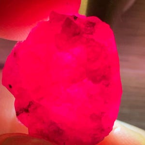 Pode incluir: Uma pedra preciosa bruta e translúcida com um tom vermelho vibrante e intenso. A pedra apresenta uma superfície irregular e facetada com texturas naturais e pequenas inclusões escuras visíveis, iluminada por trás para destacar a sua cor.