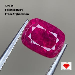 Puede incluir: Un rubí facetado de Afganistán, sujeto con pinzas. La gema es de un color rojo rosado vibrante, con un corte cojín rectangular y un patrón geométrico blanco en el centro. El texto dice "1.40 ct" y "Faceted Ruby From Afghanistan."