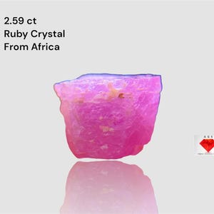 Puede incluir: Cristal de rubí rosa vibrante de África, con un peso de 2,59 ct. La gema tiene una textura rugosa y natural y se refleja debajo. También incluye el texto "Ruby Crystal From Africa".