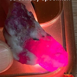Pode incluir: Um espécime de rubi de 83 ct com formato bruto e irregular, apresentando uma mistura de matriz mineral branca opaca e uma seção vibrante de cor rosa neon translúcida. O texto 83 ct Ruby Specimen está visível no topo e um logótipo AGS.