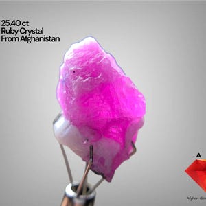 Puede incluir: Cristal de rubí rosa vibrante de Afganistán, con un peso de 25,40 ct. La gema está sujeta por pinzas metálicas sobre un fondo gris claro. La imagen incluye el texto "25.40 ct Ruby Crystal From Afghanistan".