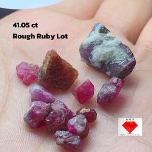 May include: A collection of rough ruby gemstones in varying shades of red and pink. The image includes the text "41.05 ct Rough Ruby Lot". The gemstones are displayed on a light-colored surface.
