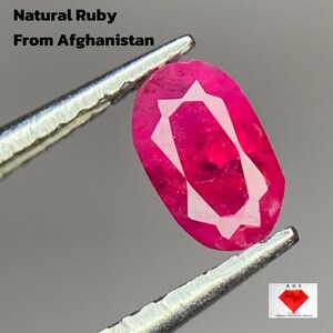 Può includere: Un rubino naturale ovale dall'Afghanistan, del peso di 0,403 ct, tenuto da una pinzetta. La gemma è di un rosso intenso con un motivo geometrico bianco al centro. L'immagine include il testo: "0,403 ct Natural Ruby From Afghanistan."