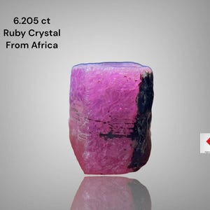 Può includere: Un vibrante cristallo di rubino rosa dall'Africa, del peso di 6,205 ct. La gemma ha una consistenza naturale ruvida con inclusioni nere scure su un lato. Il cristallo si riflette sulla superficie sottostante.