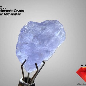 Puede incluir: Cristal de hackmanita azul claro de Afganistán, sujeto con pinzas metálicas. El cristal es de 5,80 ct y tiene una superficie rugosa y texturizada. La imagen incluye el texto "5.80 ct Hackmanite Crystal From Afghanistan".