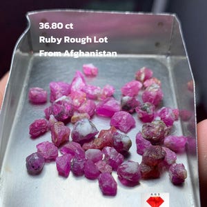 May include: A selection of rough ruby gemstones, weighing 36.80 ct, presented in a small metal tray. The rubies are various shades of pink and red, with some white and brown inclusions. Text on the tray reads "Ruby Rough Lot From Afghanistan."