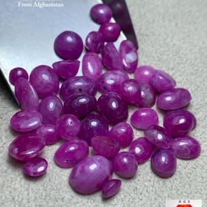 May include: A collection of deep pink ruby cabochons, with a total weight of 100 ct, are displayed on a light-colored surface. The gemstones vary in shape, including oval and round forms. Text on the image reads "Ruby Cabochons From Afghanistan."