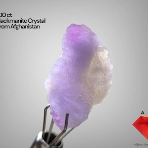 Puede incluir: Un cristal de hackmanita de 5,10 ct de Afganistán. La gema en bruto está sujeta por pinzas metálicas. El cristal es de color púrpura claro con un borde blanco. La imagen también incluye el texto "AGS" y "Afghan Gemstone Source".