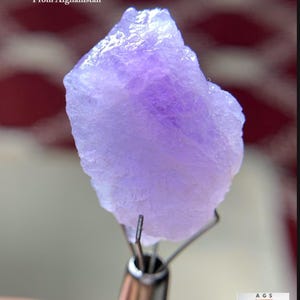 Puede incluir: Cristal de Hackmanita en bruto de color púrpura claro de Afganistán, con un peso de 17,65 ct, sostenido por una herramienta de metal. El cristal tiene una superficie rugosa y texturizada. El texto en la imagen dice "17.65 ct Hackmanite Crystal From Afghanistan."