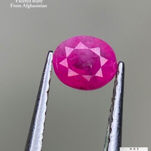 Könnte beinhalten: Ein facettierter, runder Rubin-Edelstein, gehalten von einer Pinzette. Der Rubin hat eine leuchtend rosa Farbe mit einem sternförmigen Muster, das in seine Oberfläche geschnitten ist. Der Text auf dem Bild lautet "0,34 ct Faceted Ruby From Afghanistan."