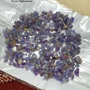 Pode incluir: Uma coleção de pedras brutas de hackmanite em tons de roxo e azul, exibida em um saco plástico transparente. O saco contém 983,70 ct de pedras do Afeganistão. A imagem também inclui um logotipo com um diamante vermelho.