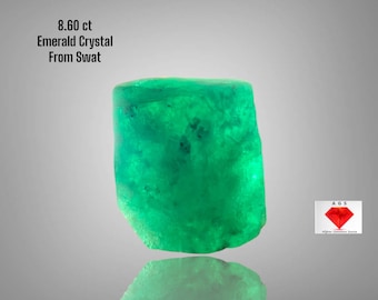 May include: A vibrant green emerald crystal from Swat, weighing 8.60 ct. The rough gemstone has a natural, irregular shape and a reflective surface. The text "8.60 ct Emerald Crystal From Swat" is visible.