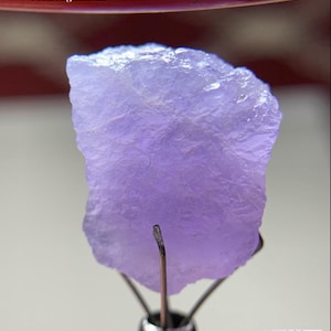 Puede incluir: Un cristal de hackmanita de color púrpura claro de Afganistán, con un peso de 20,55 ct. La piedra en bruto e irregular se sujeta con un soporte metálico. El texto "20.55 ct Hackmanite Crystal From Afghanistan" es visible.