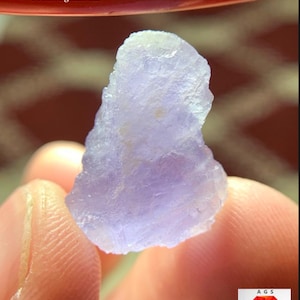 Puede incluir: Cristal de Hackmanita de color púrpura claro, tallado en bruto, de 12,20 ct, procedente de Afganistán. El cristal se sostiene entre los dedos, mostrando su forma irregular y translúcida. La imagen incluye el texto "12.20 ct Hackmanite Crystal From Afghanistan".