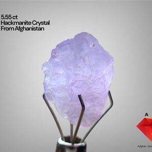 Puede incluir: Un cristal de hackmanita de color púrpura claro de Afganistán, que pesa 5,55 ct, está sujeto por un soporte metálico. El cristal tiene una superficie rugosa y texturizada. El texto de la imagen dice "5.55 ct Hackmanite Crystal From Afghanistan".