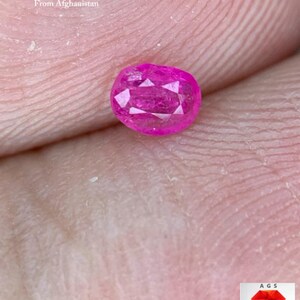 Pode incluir: Uma gema de rubi facetada em forma oval, de cor rosa vibrante. A gema é apresentada sobre um fundo de tom de pele. O texto na imagem diz "0,17 ct - Rubi facetado do Afeganistão."