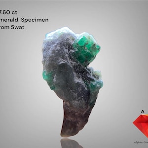 May include: A rough emerald specimen from Swat, Pakistan, weighing 27.60 ct. The stone is a mix of dark grey and green, with visible crystalline structures. The image also includes the text "Emerald Specimen From Swat".