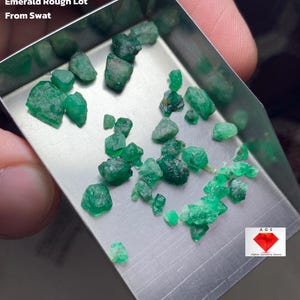 May include: A collection of rough emeralds, with a total weight of 19.75 ct, displayed in a small metal tray. The emeralds are various shades of green and have a raw, unpolished appearance. Text on the image reads "Emerald Rough Lot From Swat".