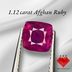 Może przedstawiać: Kwadratowy, głęboko czerwony afgański rubin o masie 1,12 karata, trzymany pęsetą. Kamień szlachetny ma biały geometryczny wzór pośrodku. U góry napis "1.12 carat Afghan Ruby". Logo "AGS Afghan Gemstone Source" znajduje się w prawym dolnym rogu.
