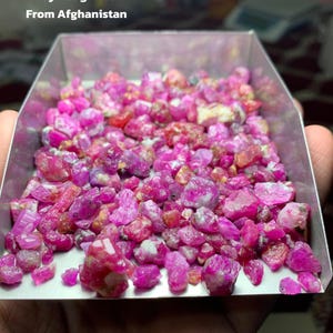 May include: A tray filled with a rough lot of rubies from Afghanistan. The gemstones are a vibrant pink and red hue, with a total weight of 536 carats. The text "Ruby Rough Lot From Afghanistan" is visible.