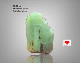 May include: A light green emerald crystal from Laghman, Afghanistan, with a weight of 38.60 ct. The rough gemstone has a translucent appearance with brown inclusions and a reflective surface. The image also includes the text "Emerald Crystal From Laghman".
