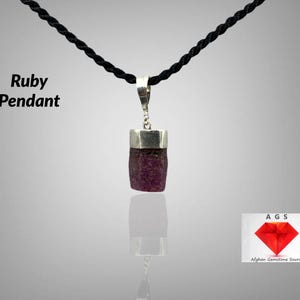 May include: A ruby pendant on a black cord. The pendant is a raw ruby set in silver. The ruby is a deep red colour and has a rough, natural texture. The pendant is labelled "Ruby Pendant".