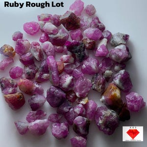 Peut inclure: Un lot de pierres précieuses de rubis brutes, de couleurs rose, rouge et marron. L'image comprend le texte "156.42 ct Ruby Rough Lot" et un logo. Les pierres précieuses varient en taille et en forme, certaines présentant des structures cristallines.