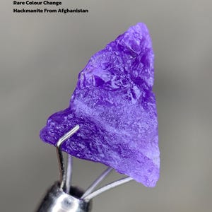 Puede incluir: Una vibrante gema de Hackmanita púrpura de Afganistán, sostenida por una herramienta de metal. La imagen incluye texto que indica que es de 4,22 ct y exhibe un raro cambio de color. La gema tiene una superficie rugosa y texturizada.