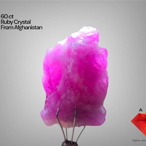 May include: A vibrant, pink ruby crystal from Afghanistan, weighing 60 ct. The gemstone is displayed on a stand against a neutral background. The image also includes the text "60 ct Ruby Crystal From Afghanistan".