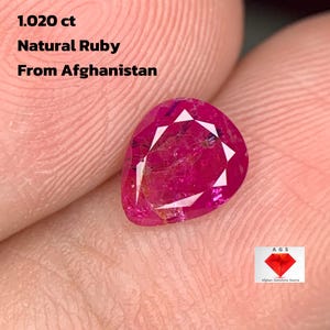 Puede incluir: Una gema de rubí natural en forma de pera de Afganistán, con un peso de 1,020 ct. El rubí es de color rojo rosado intenso con facetas que reflejan la luz. También se incluye el texto "1.020 ct Natural Ruby From Afghanistan".