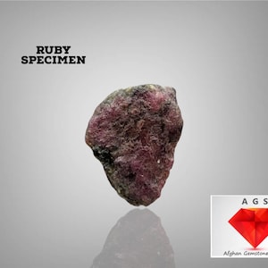 Puede incluir: Un espécimen de rubí en bruto, con colores rojo oscuro y marrón. El texto "RUBY SPECIMEN" se muestra encima de la gema. La imagen también incluye el logotipo de "Afghan Gemstone Source".