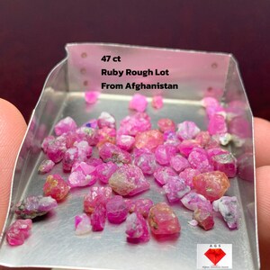 Puede incluir: Un lote de gemas de rubí en bruto, de varios tonos de rosa y rojo, exhibidas en una bandeja de metal. El texto en la bandeja dice "47 ct Ruby Rough Lot From Afghanistan."