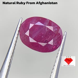 Puede incluir: Un rubí natural facetado de forma ovalada de Afganistán, sujeto con pinzas. La gema es de color rojo rosado intenso y tiene un corte brillante. El texto dice "3.00 ct Natural Ruby From Afghanistan."