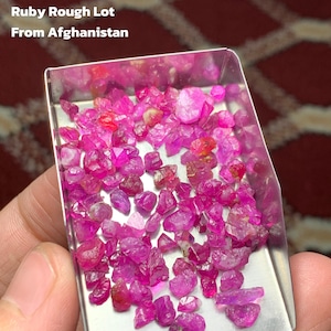 May include: A collection of rough-cut ruby gemstones in a small, rectangular metal tray. The rubies are a vibrant pinkish-red color and vary in size and shape. Text on the image reads: "67.30 ct Ruby Rough Lot From Afghanistan."