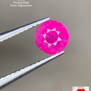 以下が含まれることがあります： ピンセットで挟まれた、丸いファセットルビー。ルビーは鮮やかなピンク色で、中央に星のような模様があります。テキストには「0.50 ct ファセットルビー、アフガニスタン産」と書かれています。