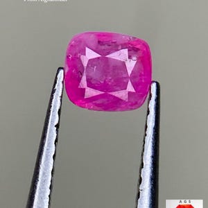 以下が含まれることがあります： ピンセットで保持されたクッションカットのルビー。宝石は鮮やかなピンク色で、内部のファセットが見えます。上部のテキストは「0.25 ct Faceted Ruby From Afghanistan」と表示されています。「AGS Afghan Gemstone Source」のロゴが右下にあります。