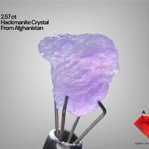Puede incluir: Un cristal de Hackmanita de color púrpura claro de Afganistán, sostenido por pinzas metálicas. El texto dice "2.57 ct Hackmanite Crystal From Afghanistan". Un logotipo de diamante rojo con el texto "Afghan Gemstone Source" está en la parte inferior derecha.