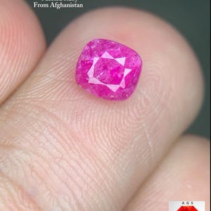 Puede incluir: Un rubí facetado, talla cojín, de aproximadamente 1,24 quilates, en un tono rosa vibrante. La gema se sostiene entre los dedos, con texto que indica su origen afgano. La gema tiene un patrón en forma de estrella en el centro.