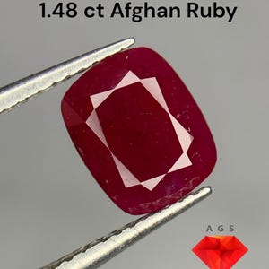 May include: A 1.48 carat red ruby gemstone, held by tweezers. The ruby is a cushion cut with a faceted design. The ruby is a vibrant red colour. The image includes the text "1.48 ct Afghan Ruby" and the logo "AGS Afghan Gemstone Source".