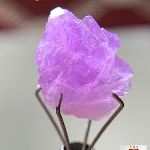 Puede incluir: Cristal de hackmanita de color púrpura claro de Afganistán, con un peso de 6,11 ct, sostenido por un soporte metálico plateado. El cristal tiene una superficie rugosa y texturizada. El fondo está borroso.