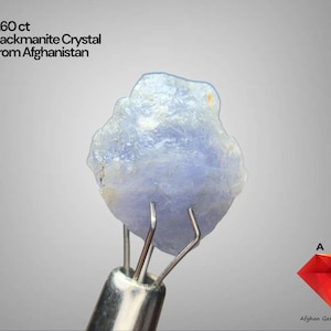 Puede incluir: Cristal de hackmanita azul claro de Afganistán, con un peso de 5,60 ct, sujeto por una herramienta de metal. El cristal tiene una forma rugosa e irregular y un aspecto translúcido. Incluye el texto "5.60 ct Hackmanite Crystal From Afghanistan".