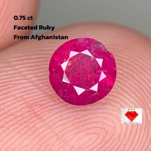 May include: A faceted ruby gemstone, 0.75 ct, with a vibrant red hue and a starburst pattern. The text "Faceted Ruby From Afghanistan" is visible. The gemstone is set against a neutral background.