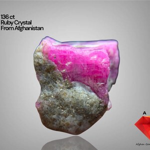 May include: A 136 ct raw ruby crystal from Afghanistan. The gemstone displays a vivid pink colour and a rough, textured surface. The image includes text that reads "Ruby Crystal From Afghanistan."