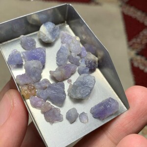 May include: A collection of rough-cut hackmanite gemstones, in shades of lavender and gray, are displayed in a small, silver metal tray. The text "40 ct Hackmanite Rough Lot From Afghanistan" is visible. The gemstones are held in a hand.