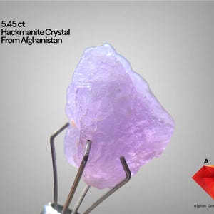 Puede incluir: Cristal de Hackmanita de color púrpura claro de Afganistán, sujeto con pinzas metálicas. El cristal pesa 5,45 quilates y tiene una forma rugosa e irregular. El texto de la imagen dice "5.45 ct Hackmanite Crystal From Afghanistan".