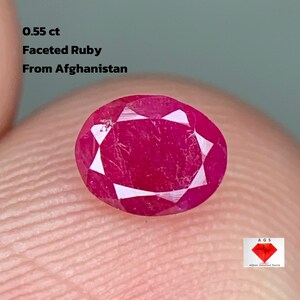 Può includere: Un rubino sfaccettato da 0,55 ct proveniente dall'Afghanistan. La gemma ovale è di un rosso intenso, con molteplici sfaccettature. L'immagine include il testo: "0,55 ct, Rubino Sfacettato, Dall'Afghanistan."