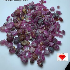 May include: A pile of rough ruby gemstones in various shades of pink, red, and brown. The image includes the text "250.45 ct Rough Ruby Lot" and a logo for Afghan Gemstone Source.